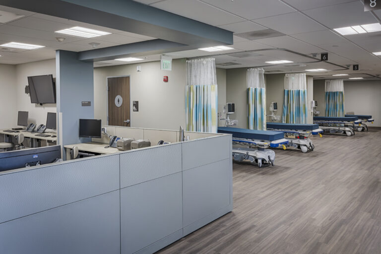 Empty hospital emergency department with cubicles, monitors, and wheeled stretchers behind privacy curtains