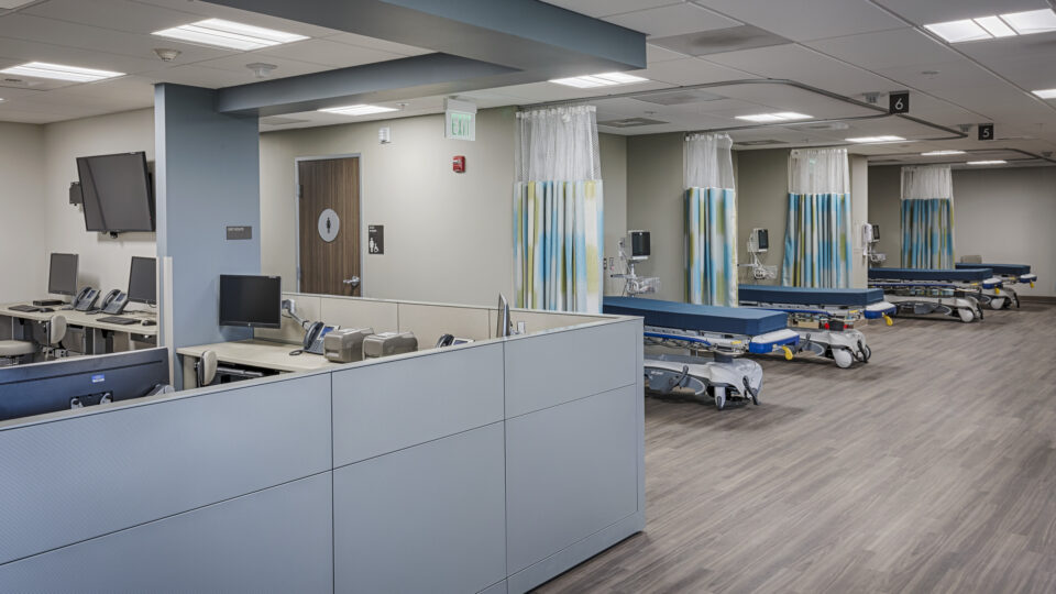 Empty hospital emergency department with cubicles, monitors, and wheeled stretchers behind privacy curtains