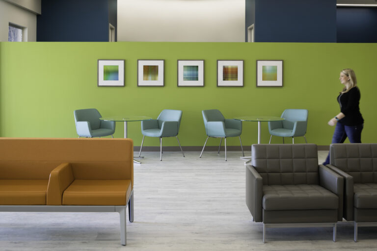 Modern lounge with colorful chairs, round tables, framed abstract art, and a woman walking past a green wall