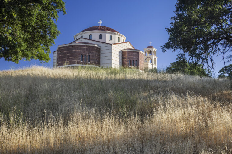 Church with domed roof and cross on a hillside, framed by trees and dry grass under a clear blue sky