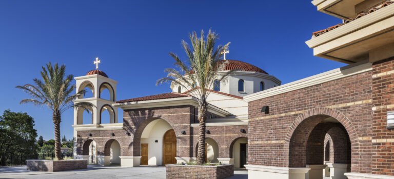 Spanish-style church exterior with brick arches, domes, palm trees, and a cross against a clear blue sky