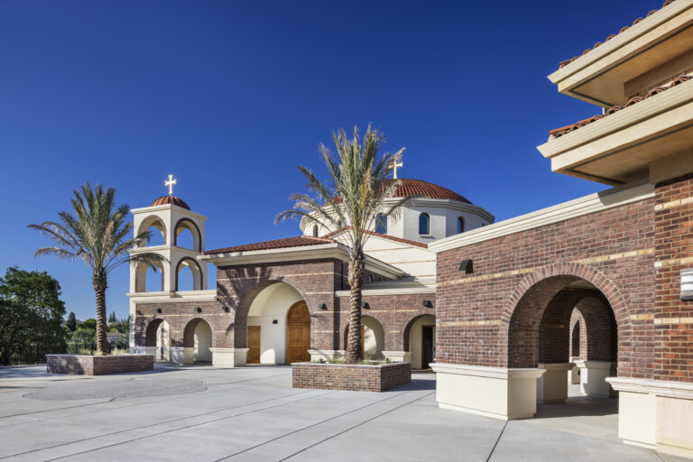 Spanish-style church exterior with brick arches, domes, palm trees, and a cross against a clear blue sky