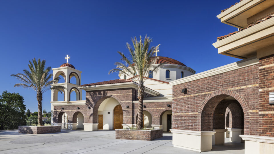 Spanish-style church exterior with brick arches, domes, palm trees, and a cross against a clear blue sky