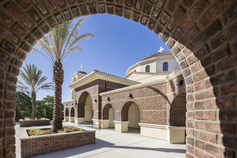 Brick arch framing a sunlit church courtyard with palm trees and domed roofs topped by crosses