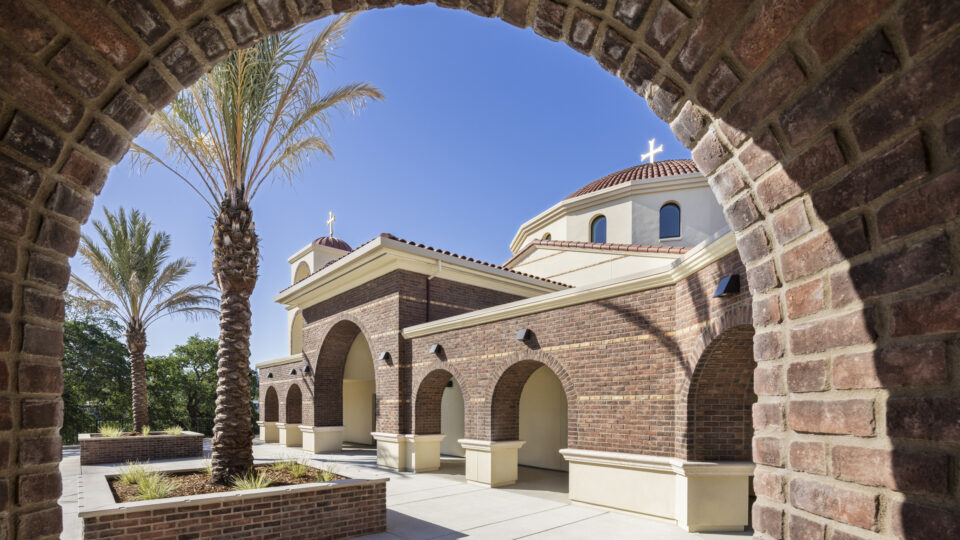 Brick arch framing a sunlit church courtyard with palm trees and domed roofs topped by crosses