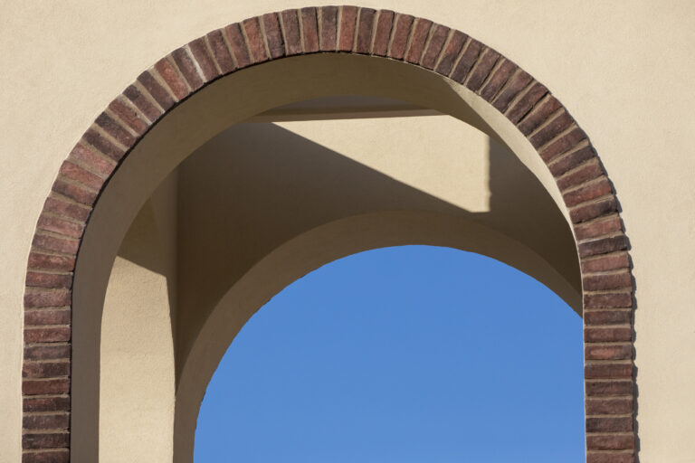 Brick-trimmed arched opening in a beige stucco wall with blue sky visible through it