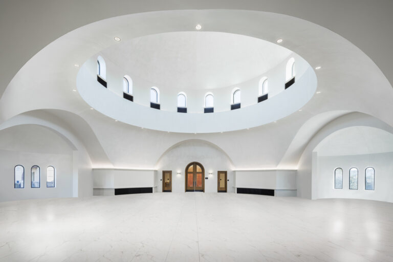 Bright white domed interior with arched windows, a central wooden door, and a large open marble floor