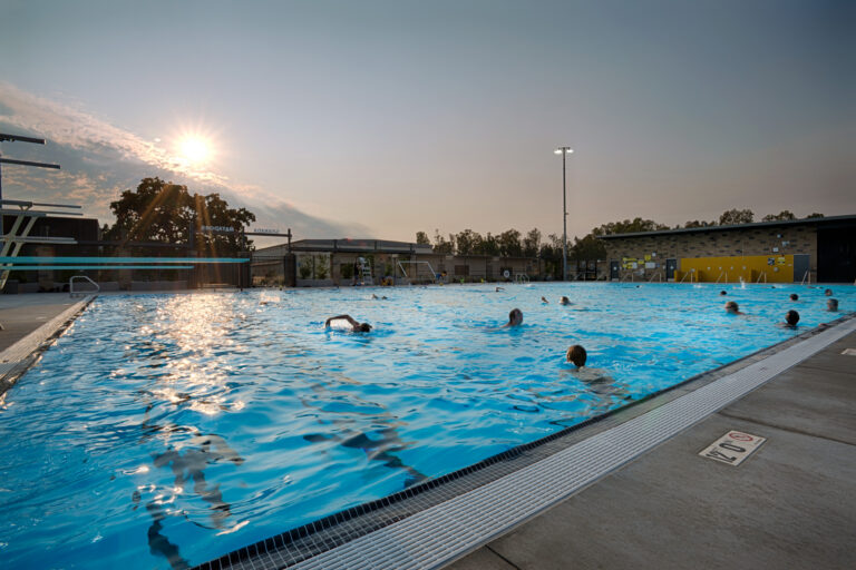 Outdoor public swimming pool at sunset with swimmers and bright sun reflections on the water