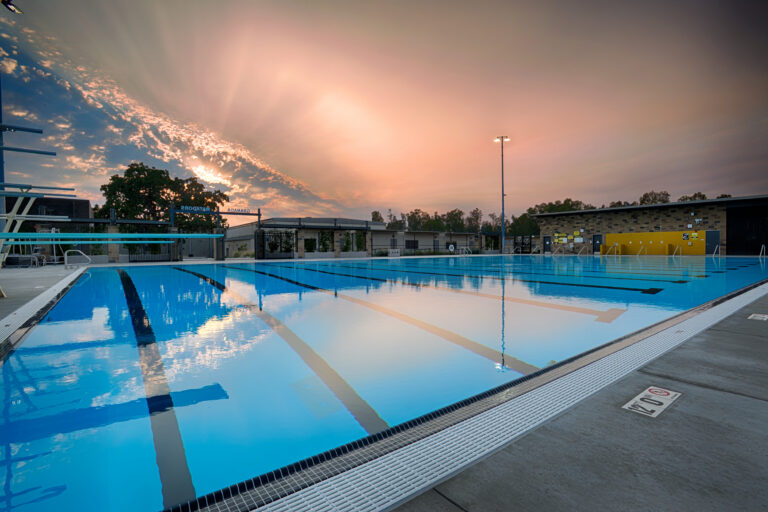 Empty outdoor lap pool at sunset with reflections on the water and a glowing sky over nearby buildings