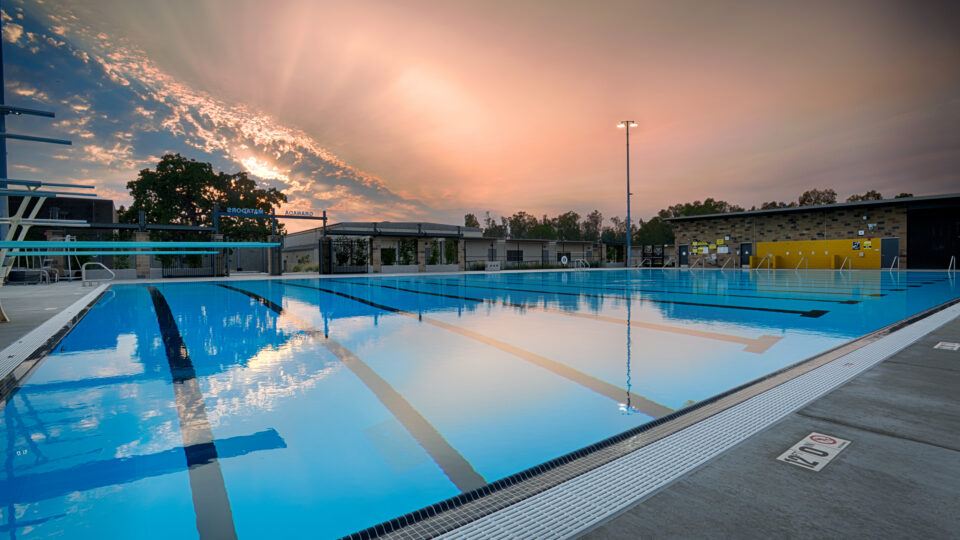 Empty outdoor lap pool at sunset with reflections on the water and a glowing sky over nearby buildings