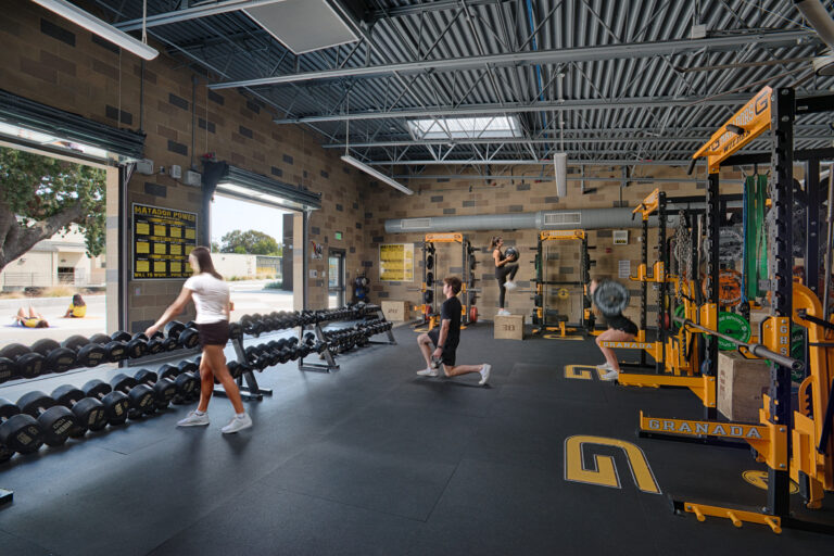 People exercising in a weight room with dumbbells, squat racks, and plyometric box jumps