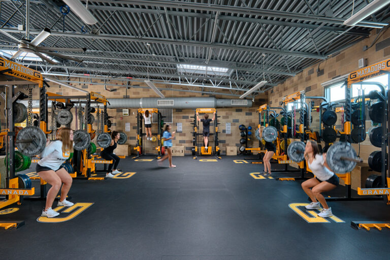 Several athletes perform barbell squats in a weight room with yellow-and-black lifting racks and the word Matadors visible