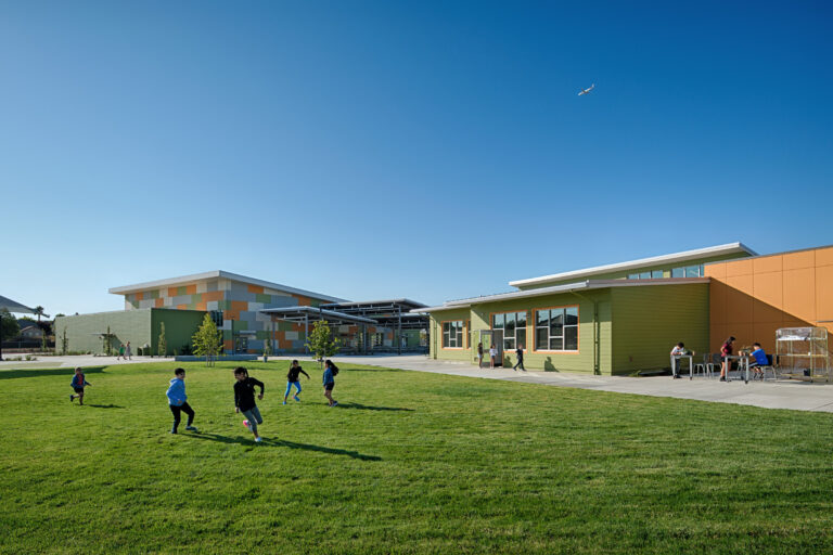 Children running on a grassy schoolyard beside colorful modern buildings under a clear blue sky, with a small airplane overhead