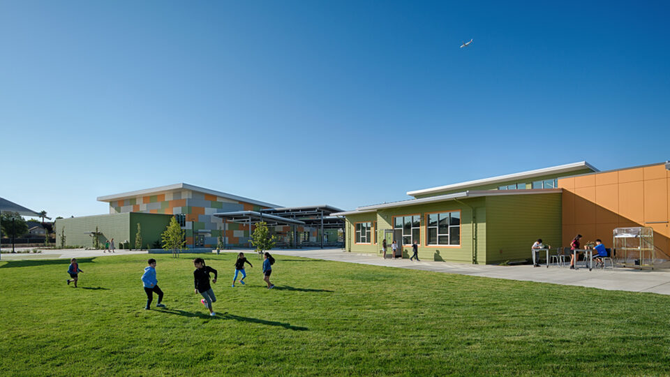 Children running on a grassy schoolyard beside colorful modern buildings under a clear blue sky, with a small airplane overhead