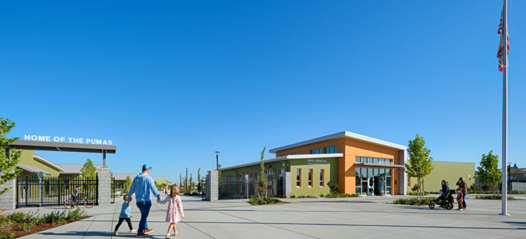 School entrance courtyard with families walking past a “Home of the Pumas” gate and a flagpole under a clear blue sky