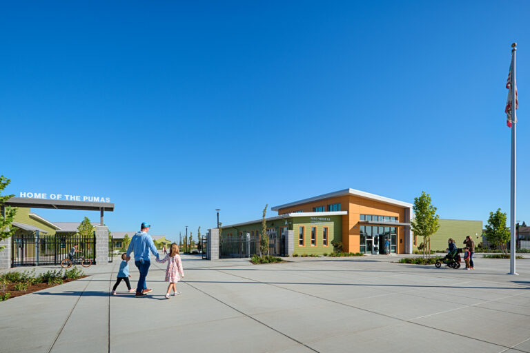 School entrance courtyard with families walking past a “Home of the Pumas” gate and a flagpole under a clear blue sky
