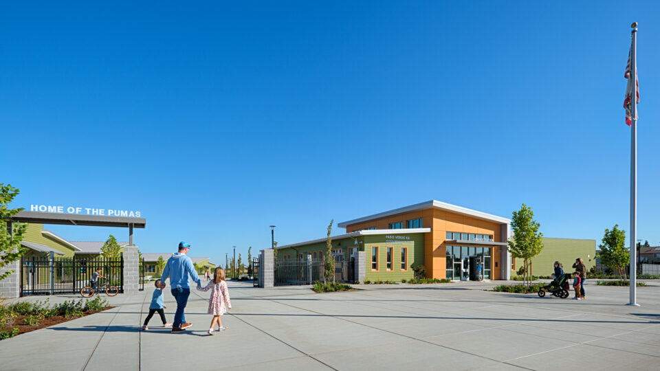 School entrance courtyard with families walking past a “Home of the Pumas” gate and a flagpole under a clear blue sky