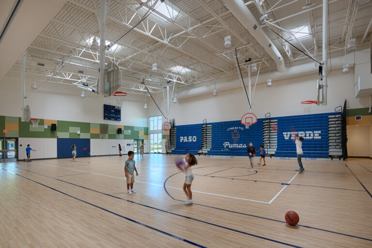 Children playing basketball in a bright indoor gym with blue bleachers and high white rafters