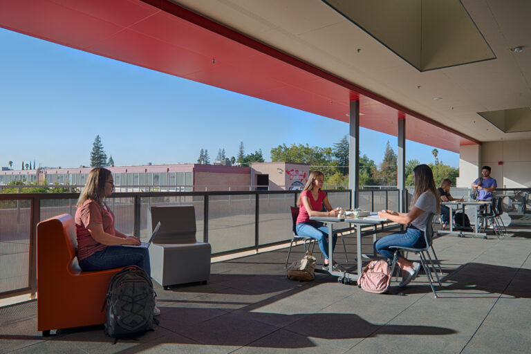 Students studying and talking on an outdoor school terrace with tables, laptops, and backpacks in bright sunlight