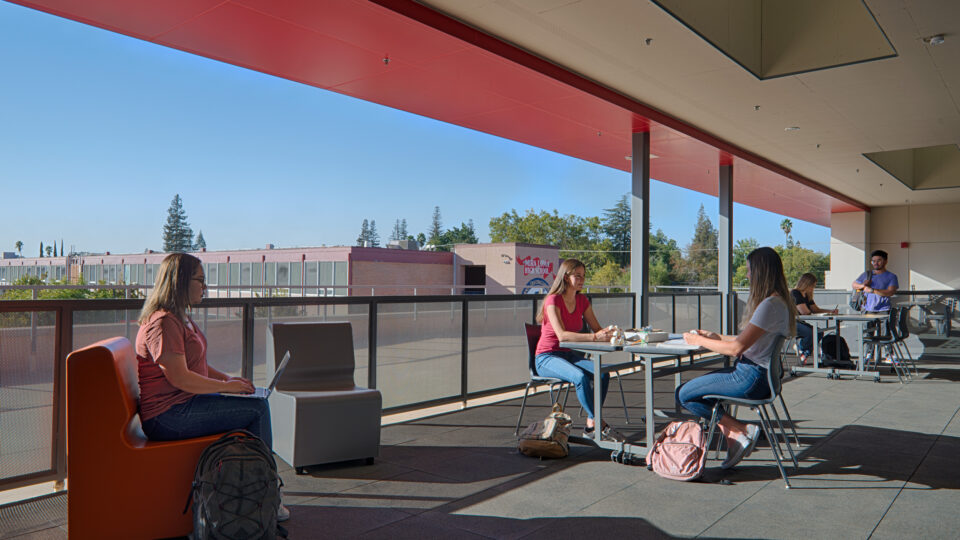 Students studying and talking on an outdoor school terrace with tables, laptops, and backpacks in bright sunlight
