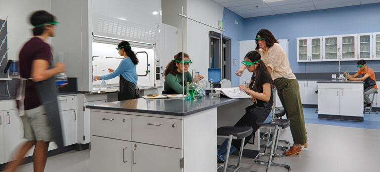 Students wearing safety goggles work together in a modern science lab around a central workstation