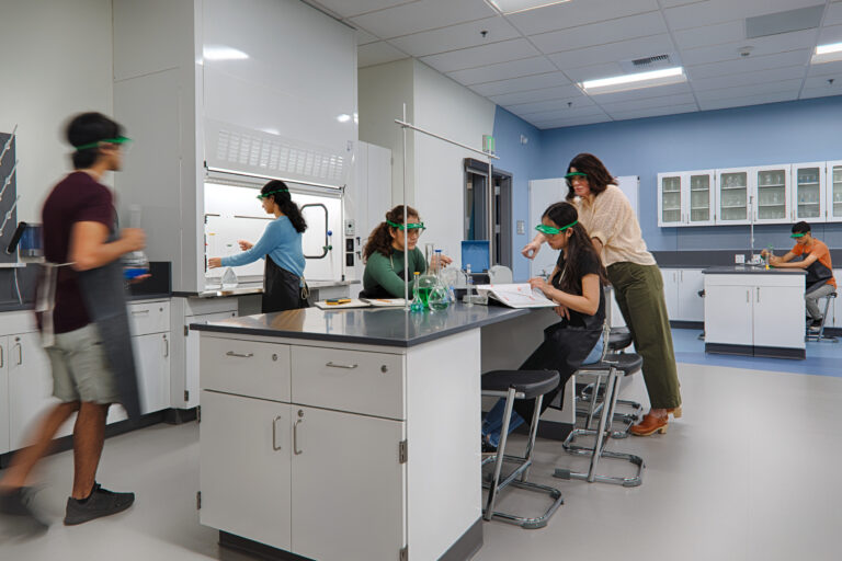 Students wearing safety goggles work together in a modern science lab around a central workstation