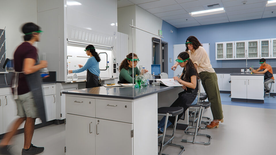 Students wearing safety goggles work together in a modern science lab around a central workstation