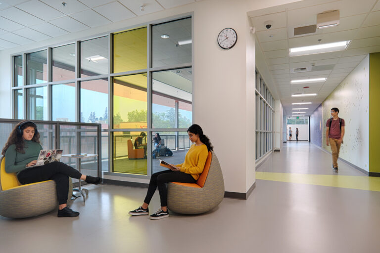 Students sitting and reading in a bright modern hallway with colorful glass walls and another student walking by