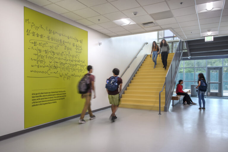Students walking through a modern stairwell beside a large green wall covered in physics equations and Einstein text