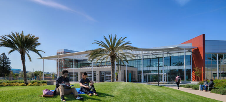 Students sitting on a grassy campus lawn in front of a modern glass building with palm trees under a clear blue sky