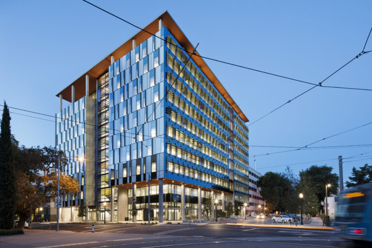 Modern glass office building at dusk with lit windows, an orange roof, and trolley wires overhead