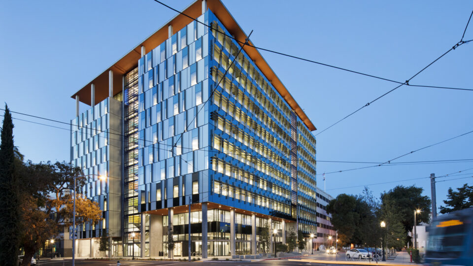 Modern glass office building at dusk with lit windows, an orange roof, and trolley wires overhead