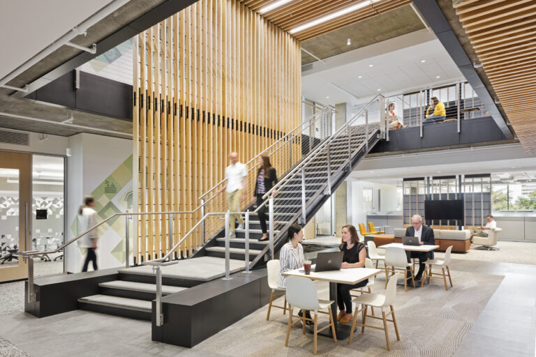 Modern office atrium with a central staircase, wood slat screens, and people working and talking at tables