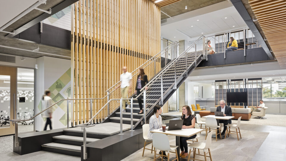 Modern office atrium with a central staircase, wood slat screens, and people working and talking at tables