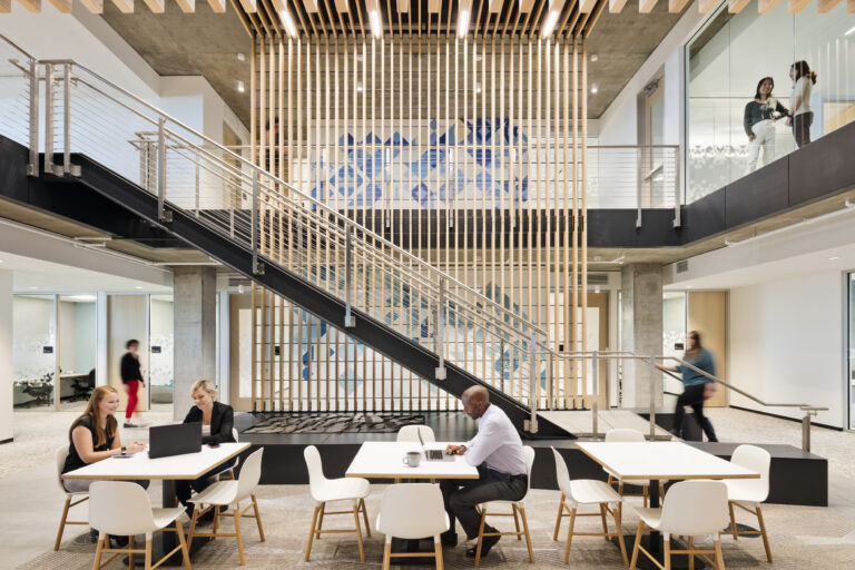 Modern office atrium with stairs, glass-walled meeting rooms, and people working at tables below