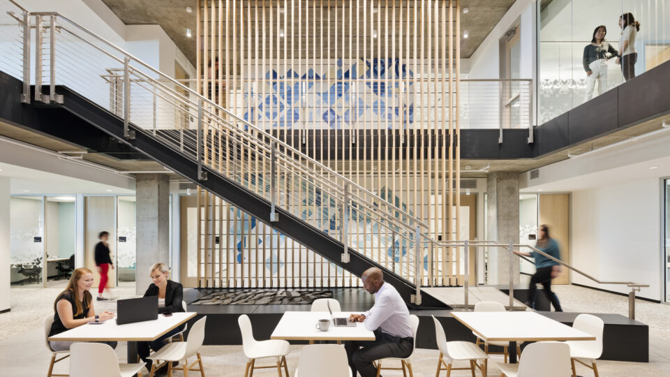 Modern office atrium with stairs, glass-walled meeting rooms, and people working at tables below