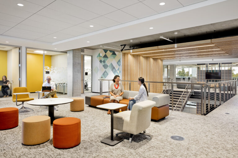 Modern open-plan office lounge with people working and talking among colorful chairs, tables, and sofas