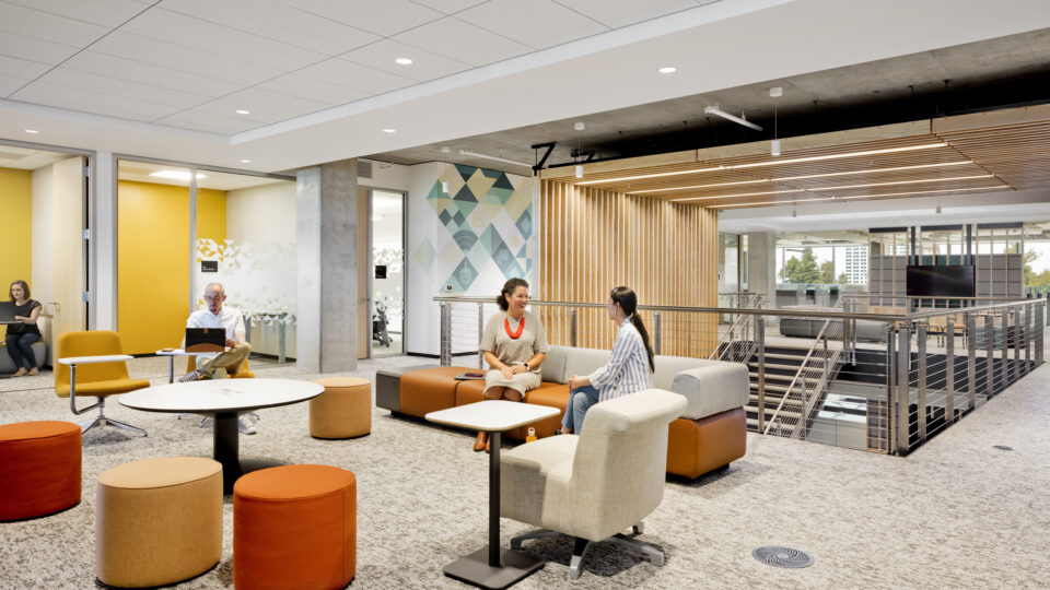 Modern open-plan office lounge with people working and talking among colorful chairs, tables, and sofas