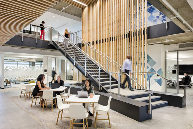 Modern office lounge with people working at tables beside a staircase and wood-slat divider
