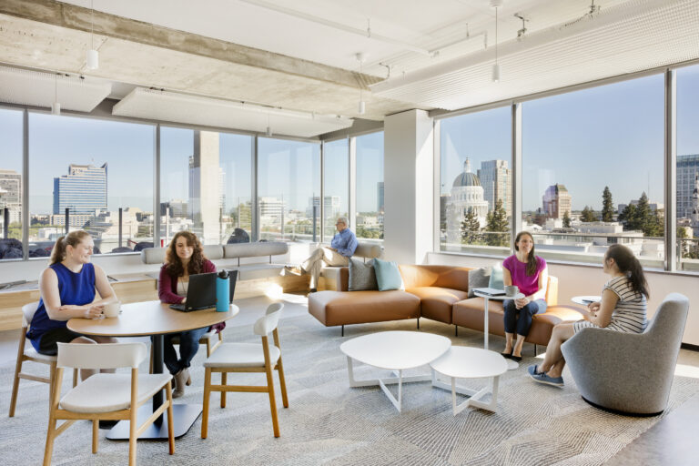 Bright modern office lounge with people chatting and working by large windows overlooking a city skyline