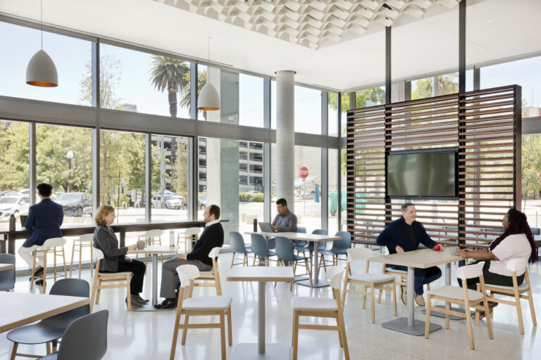 Bright modern cafe with people sitting at tables by large windows and a TV mounted on a slatted wood divider