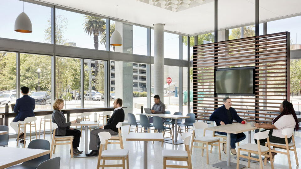 Bright modern cafe with people sitting at tables by large windows and a TV mounted on a slatted wood divider