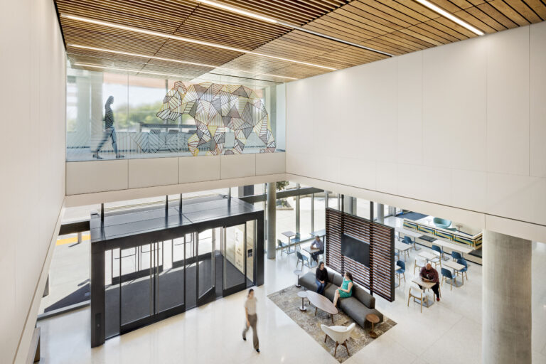 Modern office lobby with glass walls, wood slat ceiling, lounge seating, and a geometric bear mural overhead