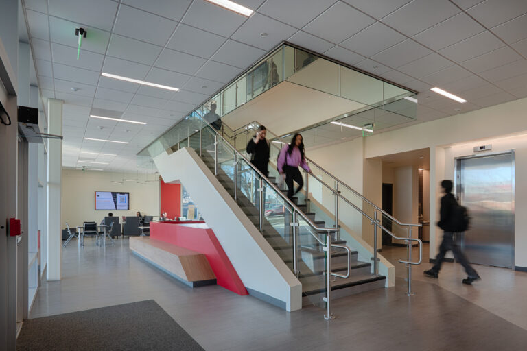 Modern lobby with a glass staircase, elevator, and people walking through a bright open office space