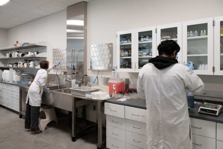 Two lab workers in white coats and gloves prepare samples at a stainless-steel laboratory sink and bench