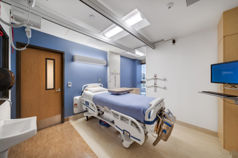 Modern hospital patient room with an empty bed, overhead rails, monitor, and blue accent wall