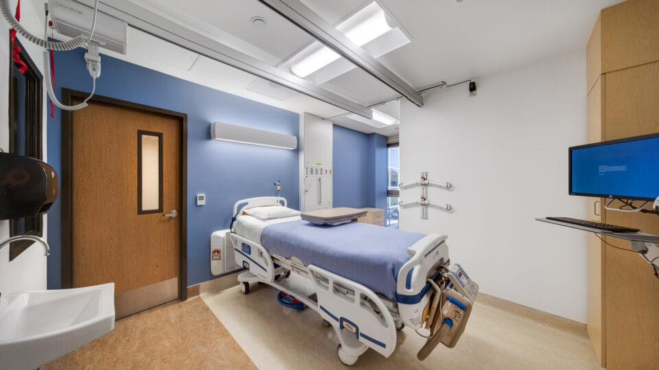 Modern hospital patient room with an empty bed, overhead rails, monitor, and blue accent wall