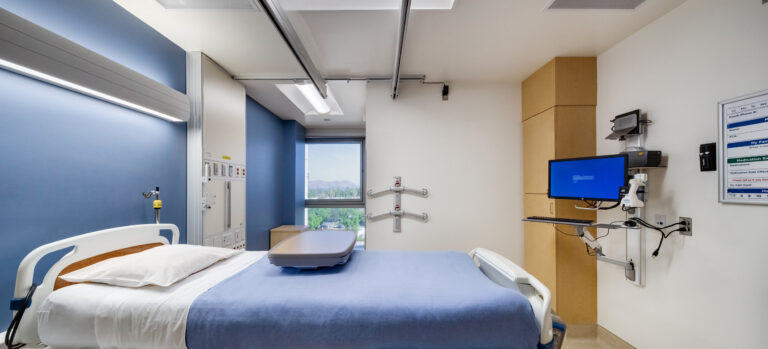 Empty hospital patient room with an adjustable bed, medical monitors, and a window view outside