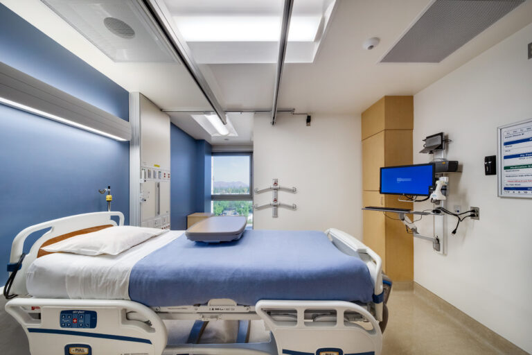 Empty hospital patient room with an adjustable bed, medical monitors, and a window view outside