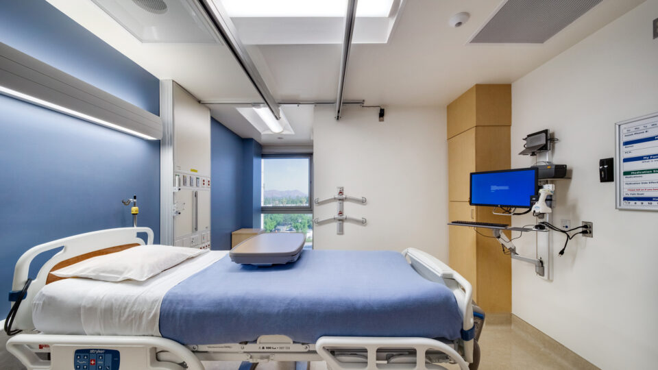 Empty hospital patient room with an adjustable bed, medical monitors, and a window view outside